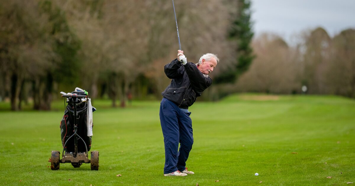 A senior golfer at the top of a swing on a UK parkland course, trolley beside him