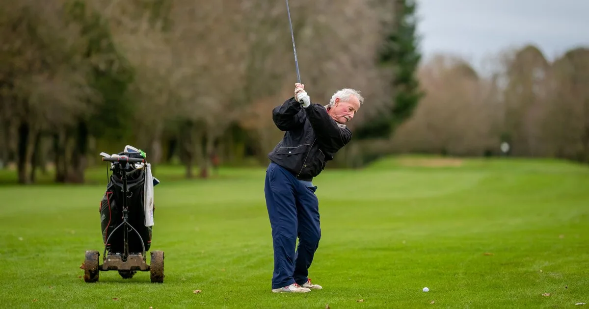 A senior golfer at the top of a swing on a UK parkland course, trolley beside him