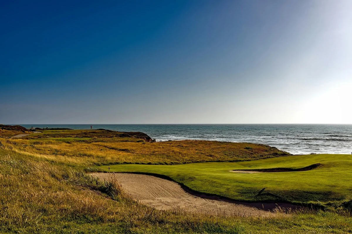A golfer playing a shot on an exposed links course with a clear sky and strong wind